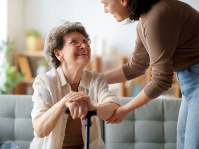 A woman helps her aging mother sit down on the couch, acting as a family caregiver.