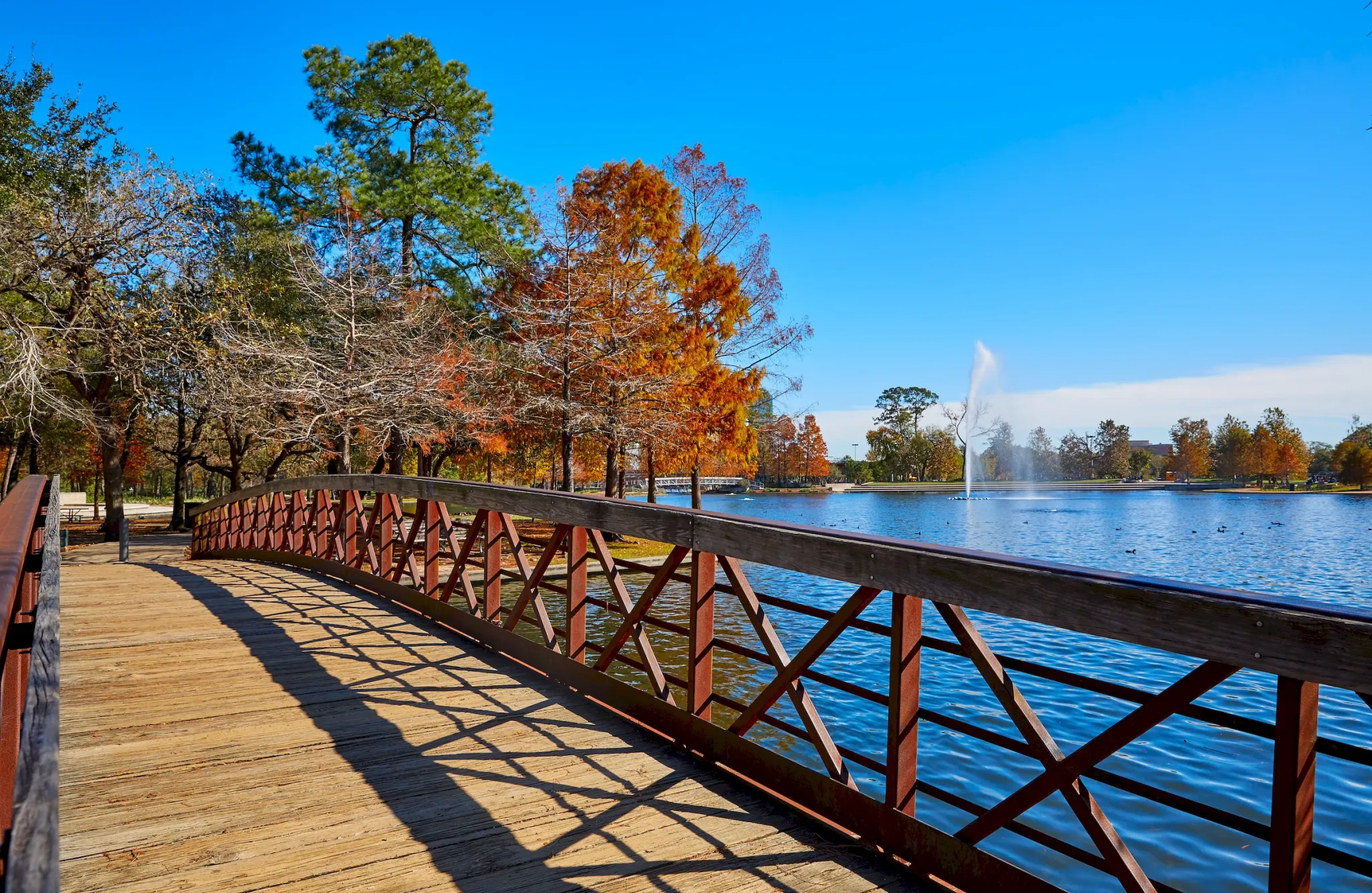 Wooden bridge over a lake with autumn trees, clear sky, and a water fountain in the distance.