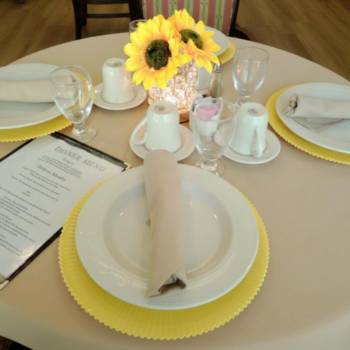 Round table set for four at Tarina of Kingwood with yellow chargers, beige napkins, a menu, and a sunflower centerpiece.