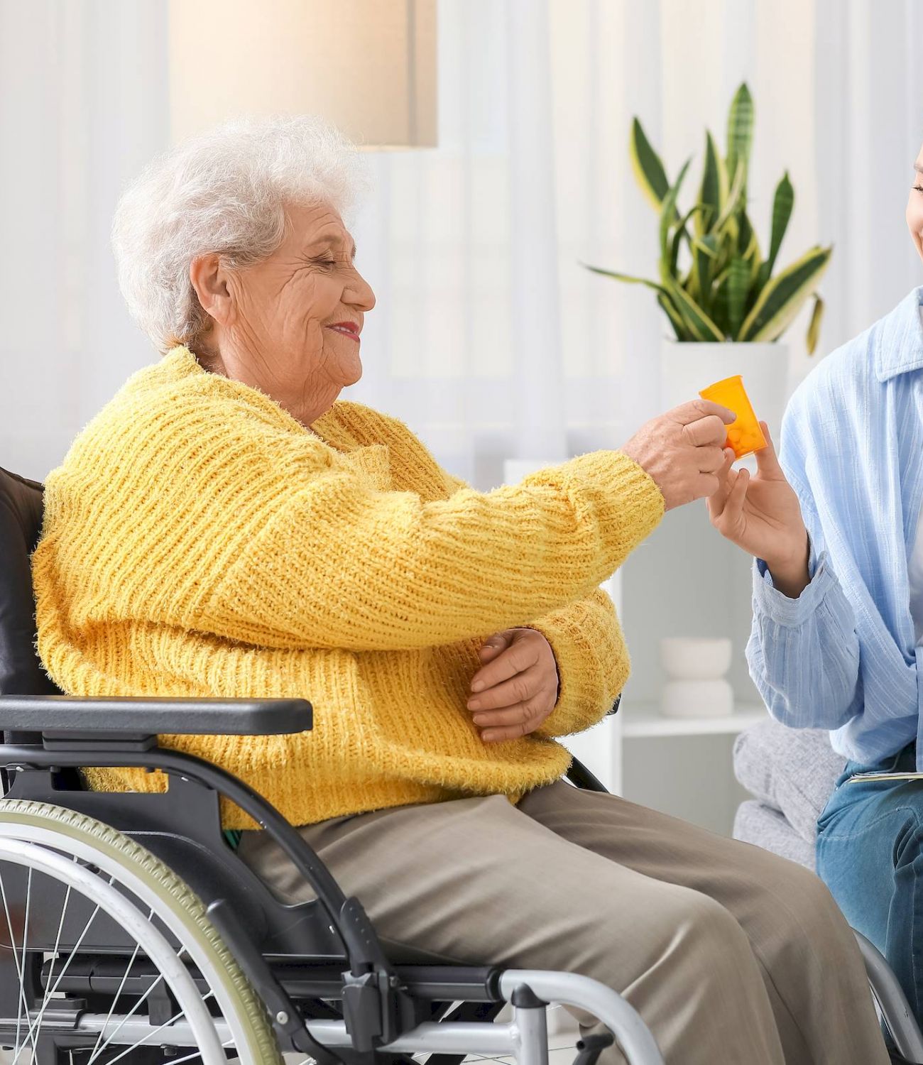 Young woman smiles as she hands a card to Tarina of Stockton, an elderly woman in a yellow sweater, seated indoors in her wheelchair.