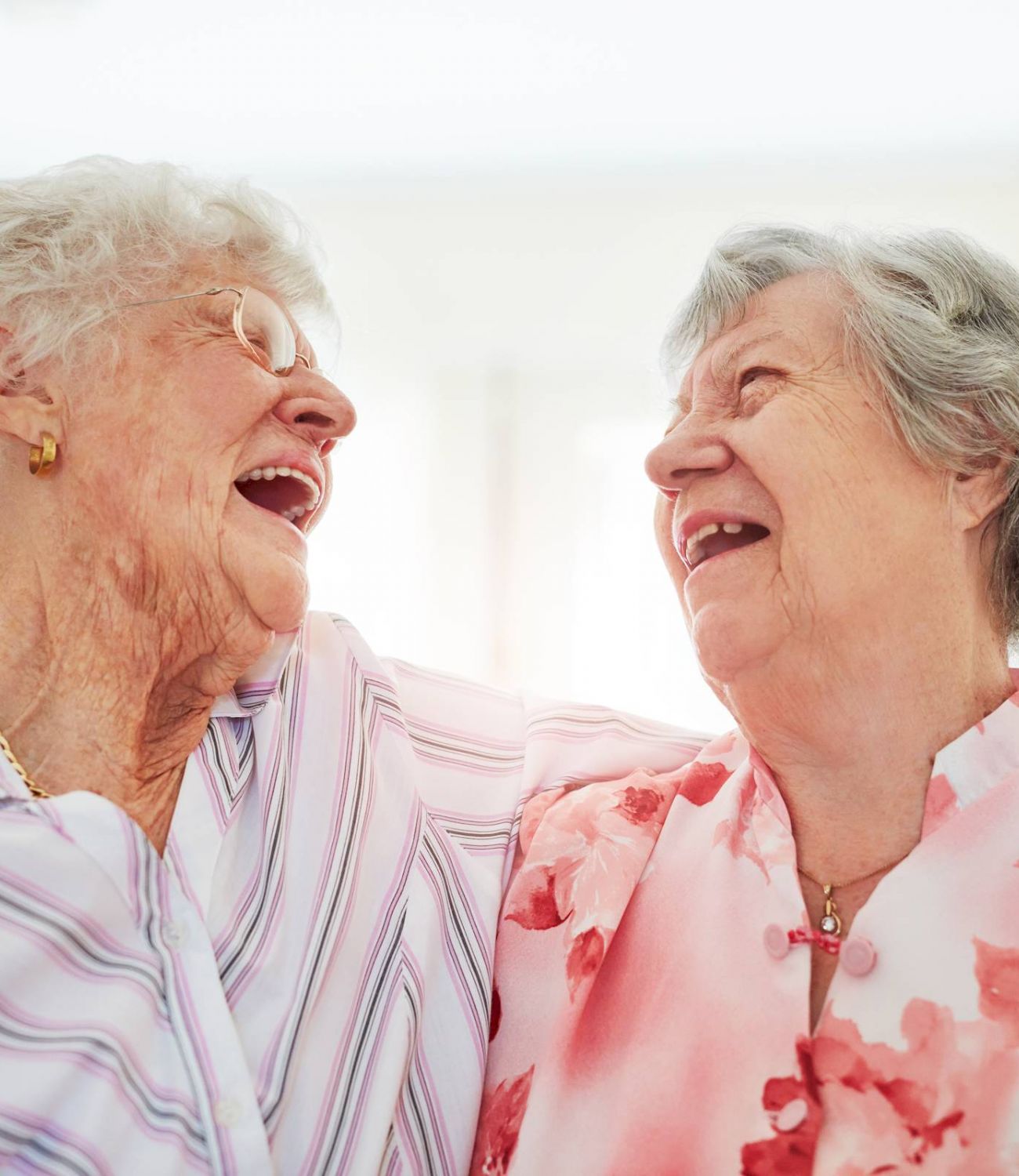 Two elderly women smiling and laughing together, arms around each other in a bright room.