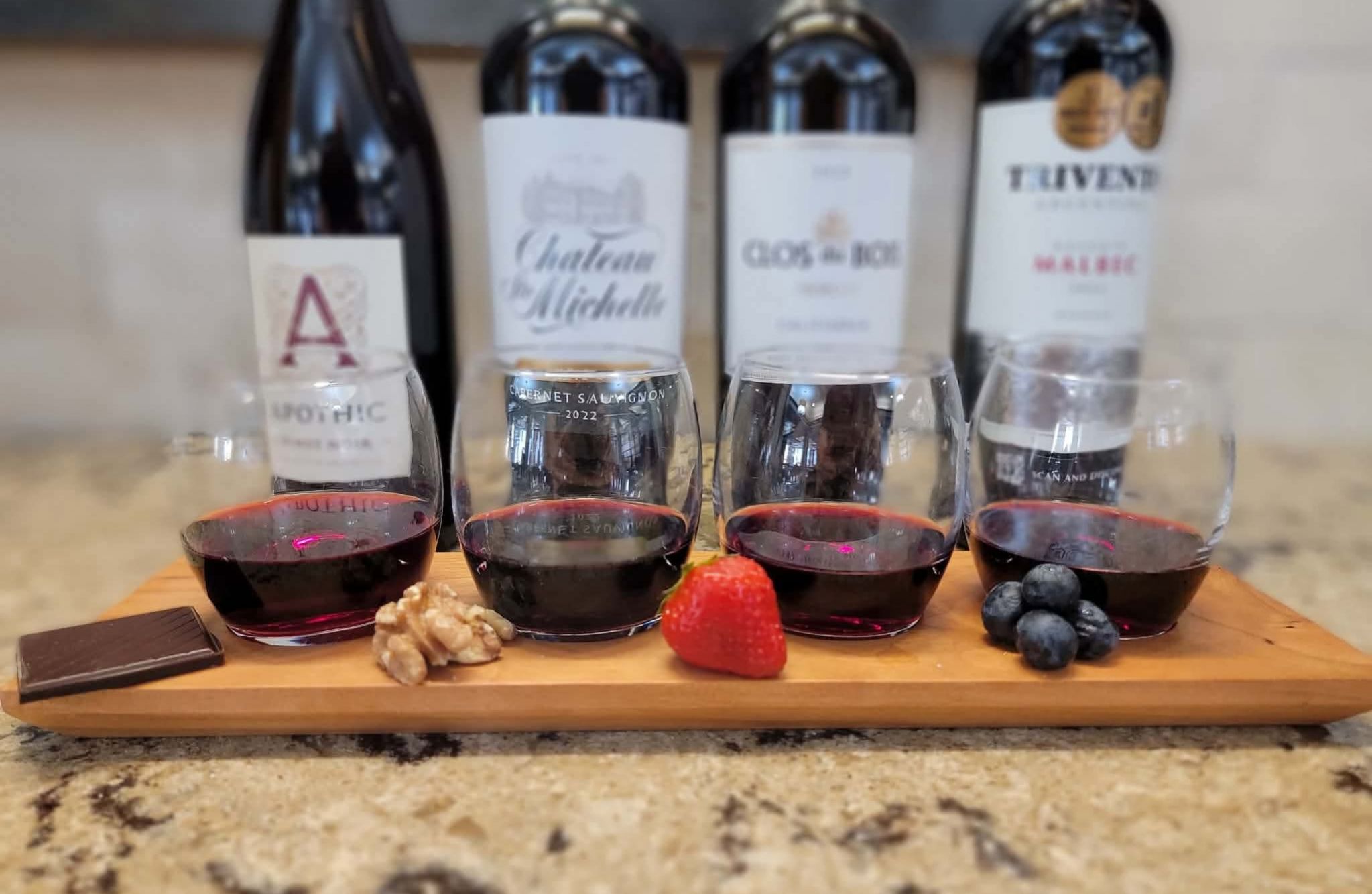 Four glasses of red wine with chocolate, walnut, strawberry, and blueberries—a tasteful touch to living well—on a wooden tray with bottles behind.
