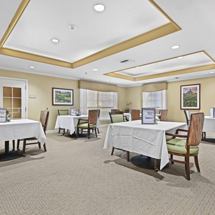 A bright dining room at Tarina of Cypresswood features tables for four, white tablecloths, and wicker chairs in a senior living facility.