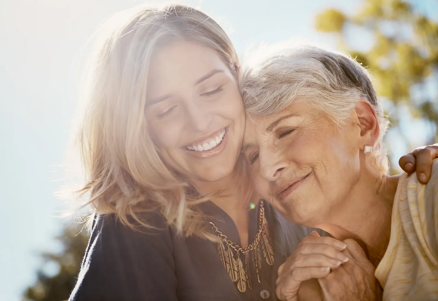 Two women, one younger and one older, smiling and hugging outdoors in warm sunlight.