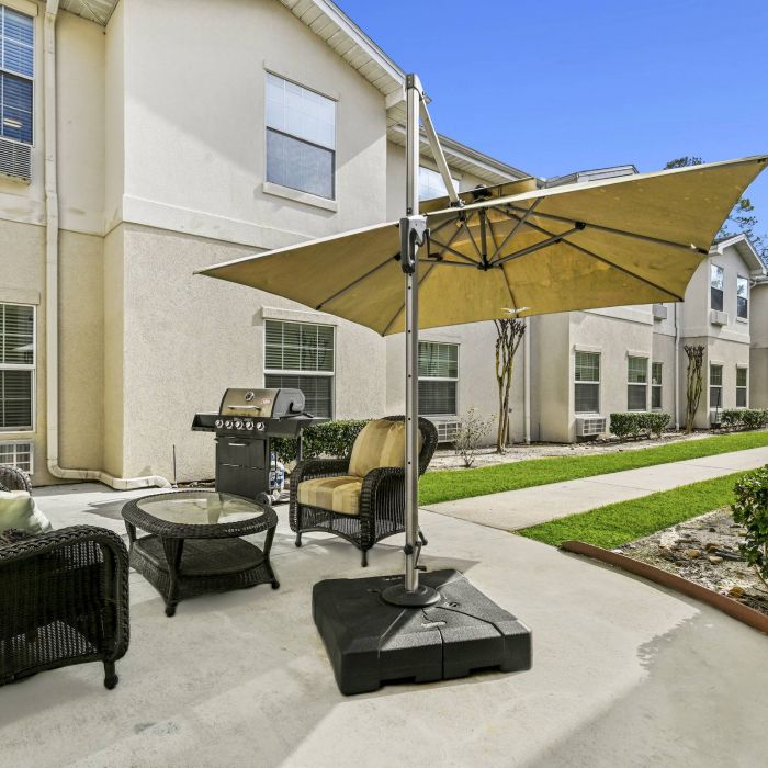 Outdoor patio at Tarina of Cypresswood features wicker seating, a glass table, grill, and large umbrella beside the walkway and garden.