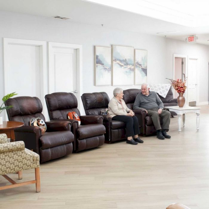 Two elderly people sit on brown recliners in a bright, modern room with minimalist decor and paintings on the wall.