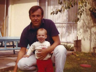 An adult crouches, hugging a smiling baby in a yard—Honoring Every Story amid patio furniture and the home's warm background.