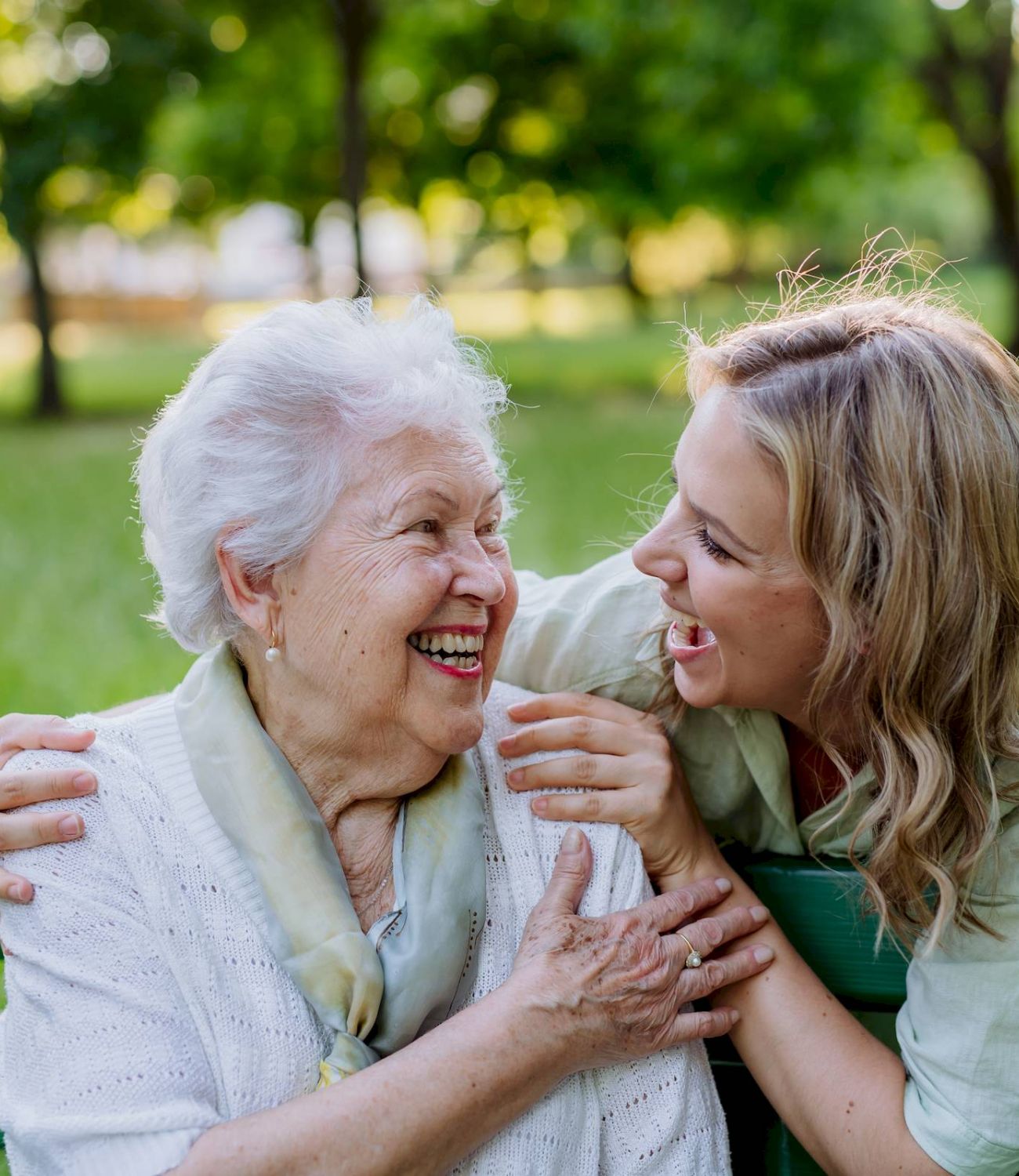 Elderly woman and younger woman laughing together on a park bench, sharing a joyful moment.