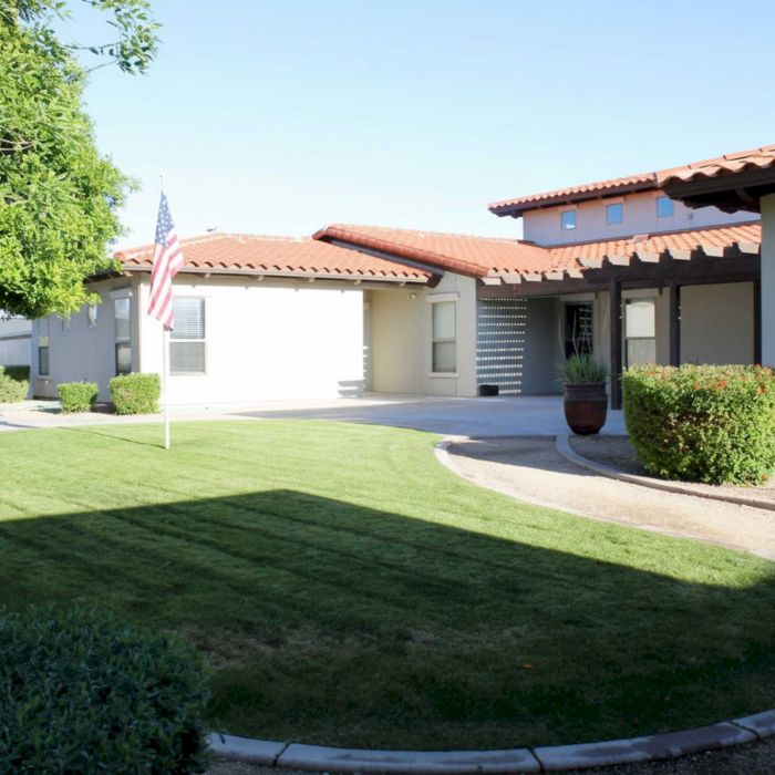 Single-story house with red-tiled roof, flagpole, lawn, shrubs, and a curved sidewalk in front.