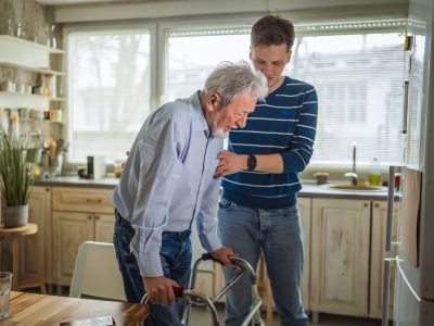 Young man helps elderly man with a walker in a bright kitchen, discussing signs of assisted living and future care options.