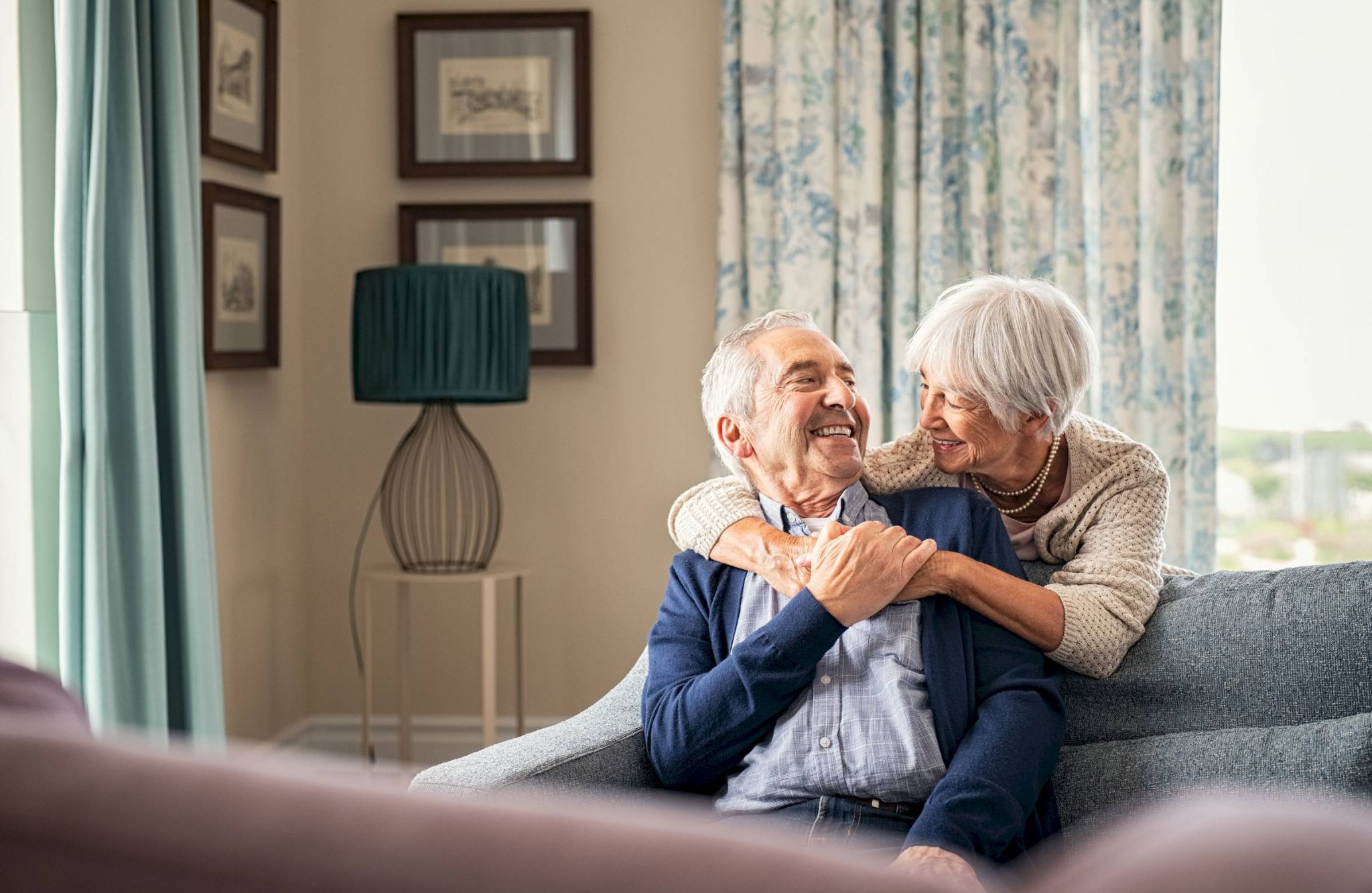 Senior couple sitting on a couch, smiling and embracing each other in a cozy, sunlit living room.