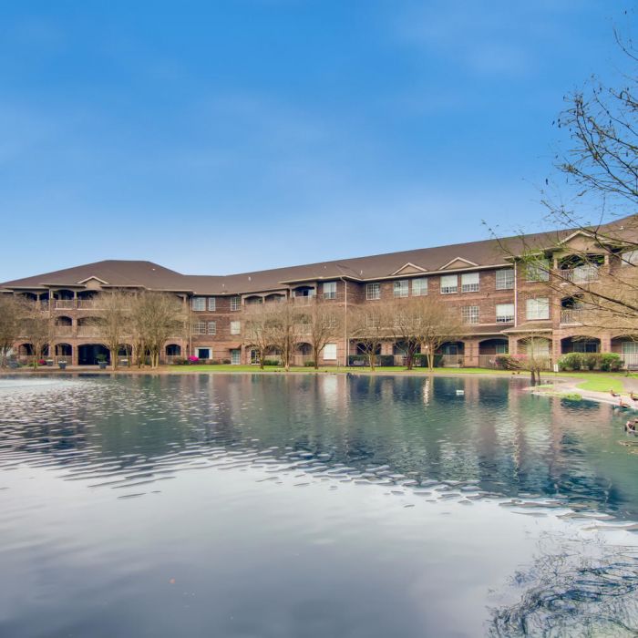 The Terraces at Kingwood, TX with a Large pond with a water fountain in front of The Terraces at Kingwood, surrounded by leafless trees under a blue sky.