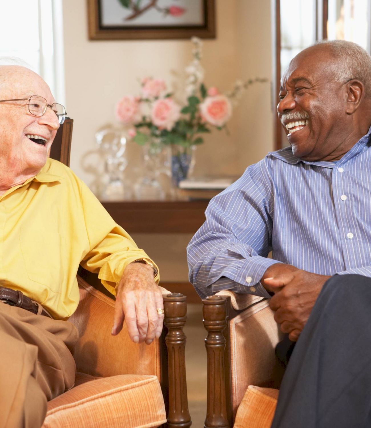 Two elderly men sit in armchairs, smiling and laughing together in the cozy, well-lit living room at Tarina of Kingwood.