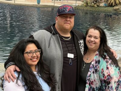 Three people smiling and posing together by a pond, Honoring Every Story and Remembering Ours amid lush greenery.