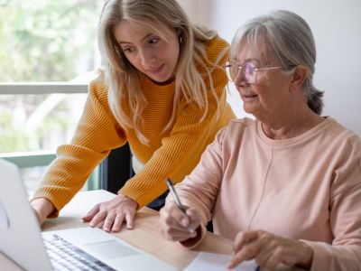 Older woman writes notes while younger woman helps her use a laptop at a table in a bright room.