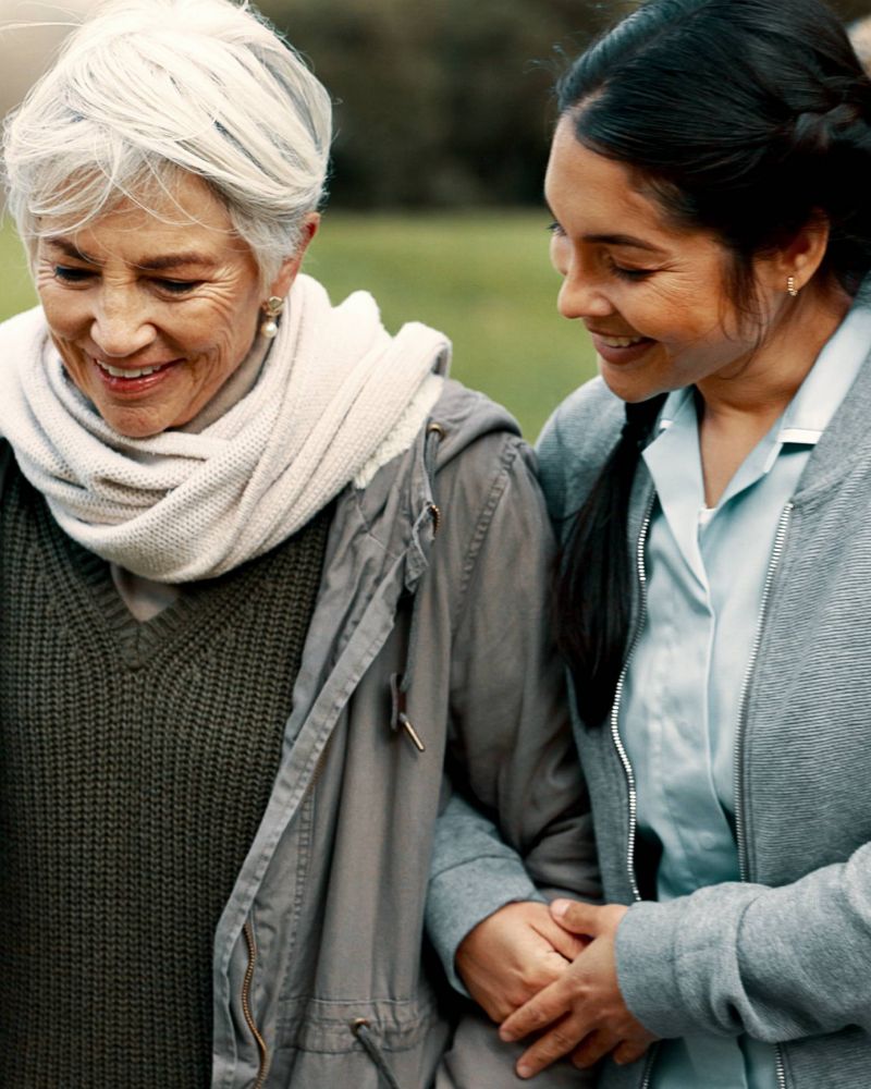 A younger woman smiles as she supports an older woman walking outdoors on a grassy path.