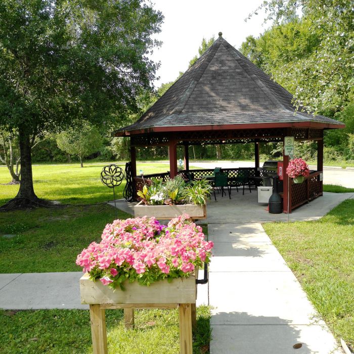 A wooden gazebo with chairs at Tarina of Kingwood, surrounded by greenery and pink flowers in planters along a sidewalk.