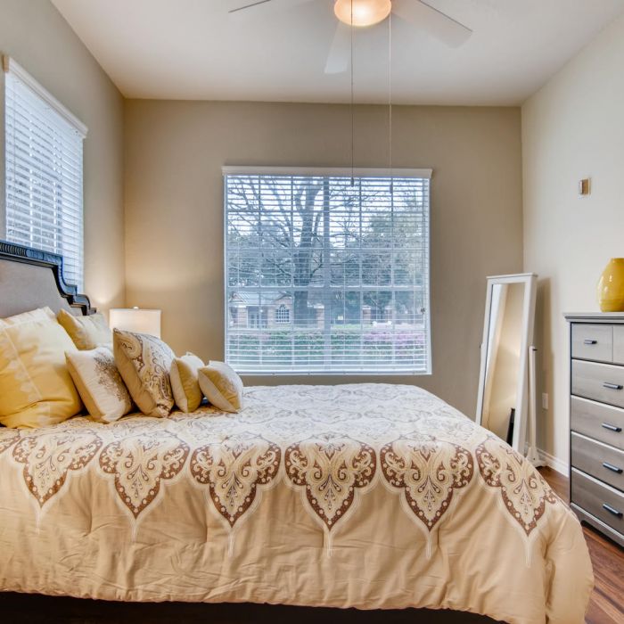 Cozy bedroom with a patterned bed, yellow pillows, dresser, mirror, and large window at Terraces at Kingwood senior living.