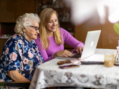 A young woman helps an elderly woman use a laptop at a table in an assisted living setting, both smiling and engaged.