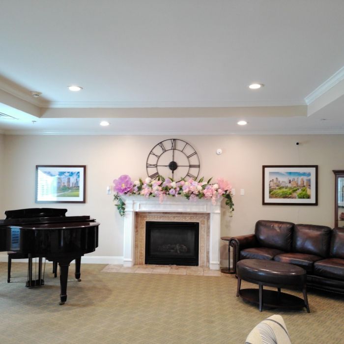 A living room at Tarina of Kingwood features a grand piano, leather sofa, fireplace, large clock, and floral mantel decor.
