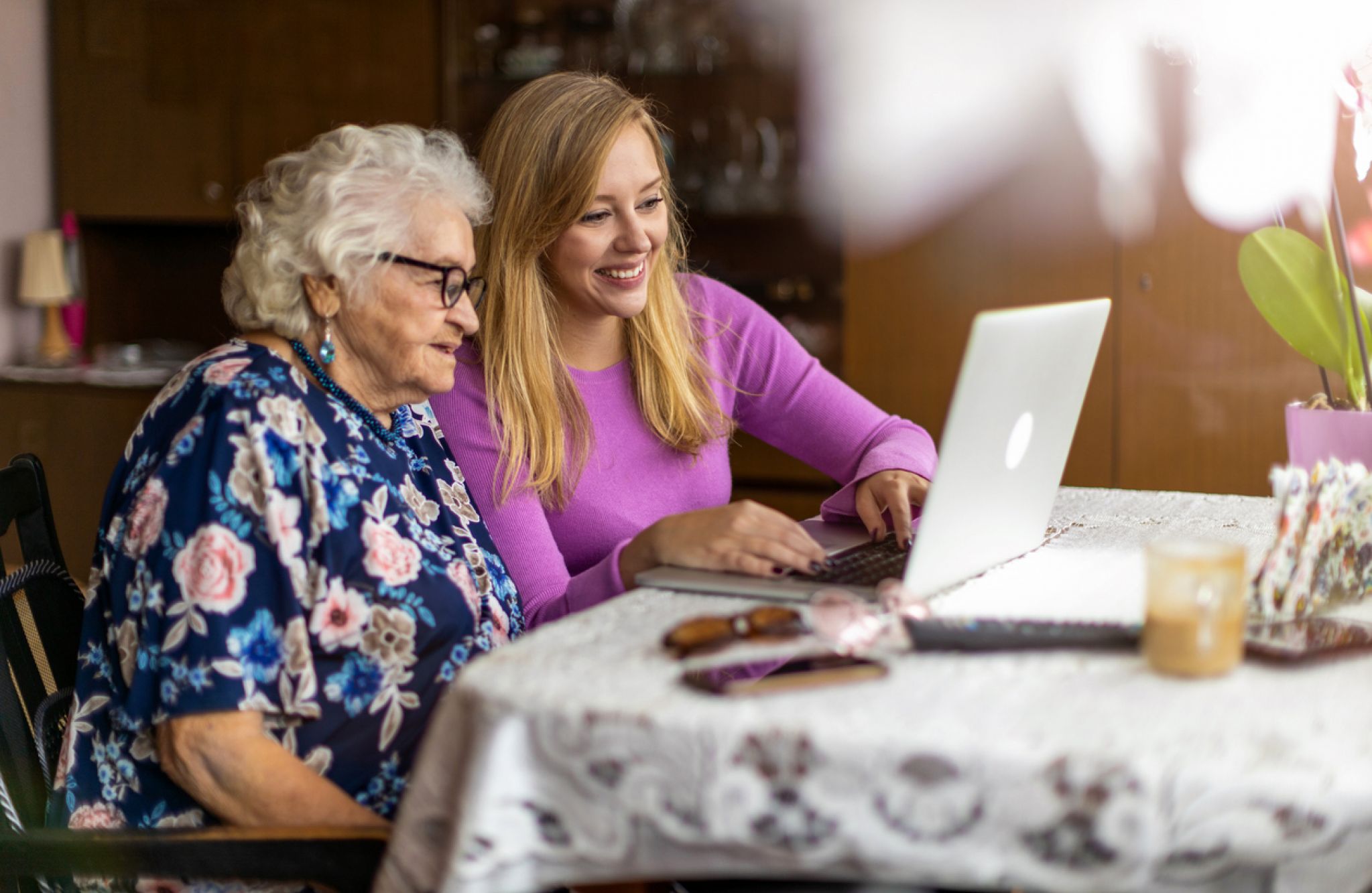 A young woman helps an elderly woman use a laptop at a table in an assisted living setting, both smiling and engaged.