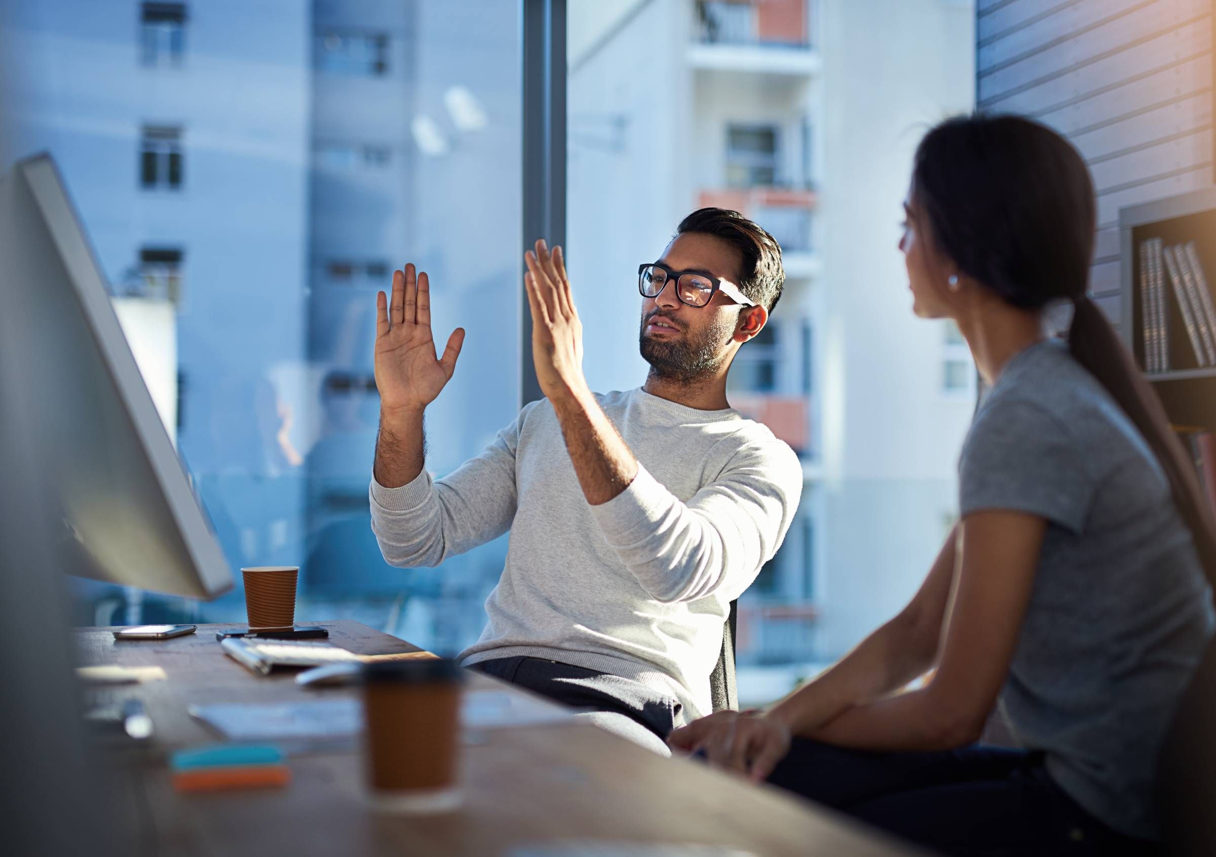 Man gesturing while explaining something to a woman in an office with a computer and large window.