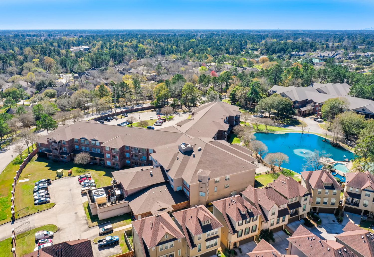 Aerial view of The Terraces at Kingwood, a senior living community with a pond, trees, and parked cars on a sunny day.