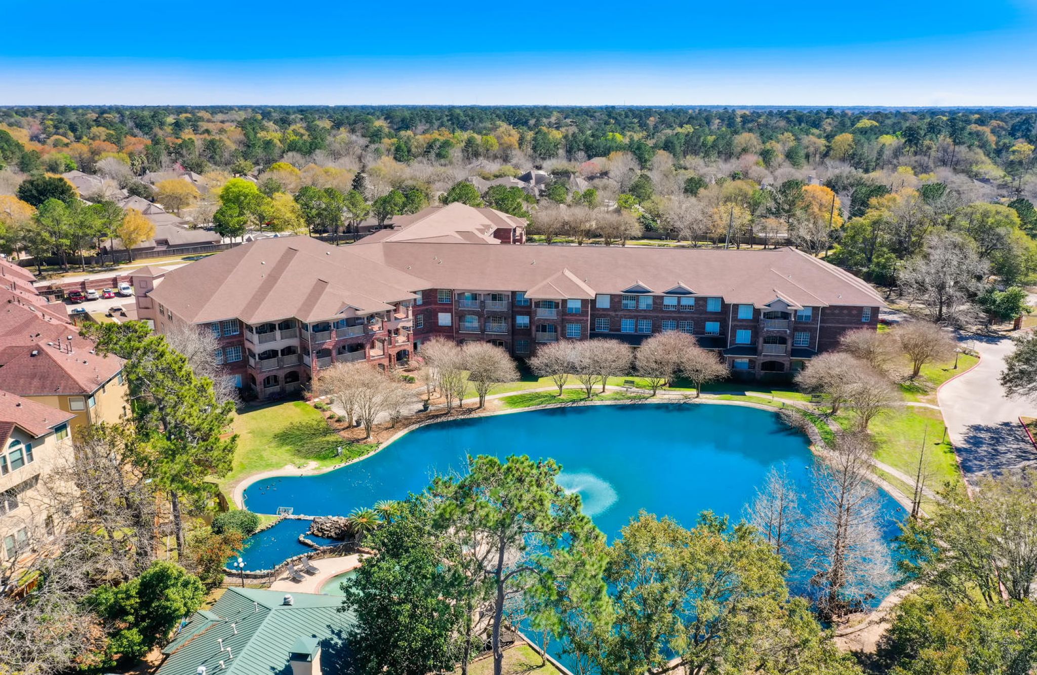 The Terraces at Kingwood, TX with an Aerial view of The Terraces at Kingwood, a large building by a pond amid trees and greenery under a clear blue sky.