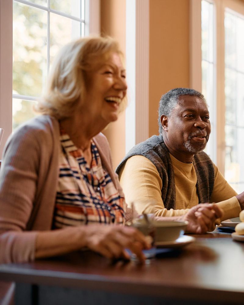 Four older adults laughing and talking together around a dining table in a brightly lit room.