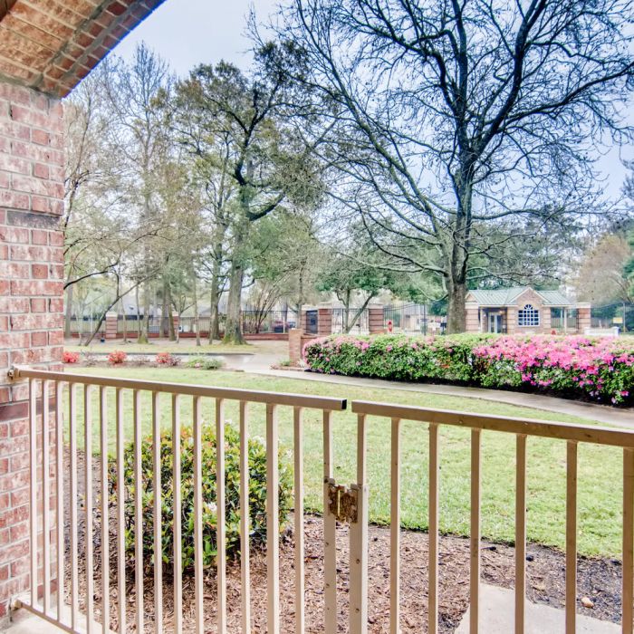 Enjoy a relaxing view from a brick porch at The Terraces at Kingwood, overlooking blooming bushes and trees in a peaceful neighborhood.