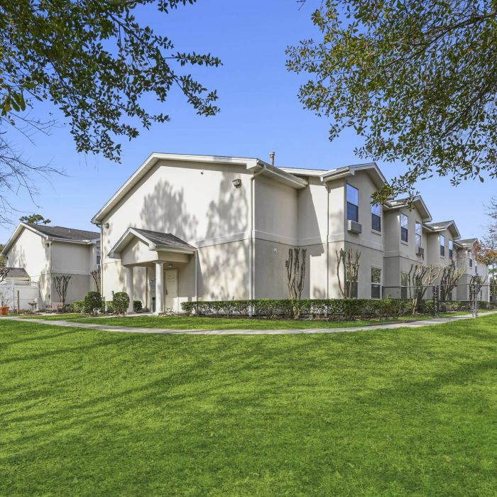 Tarina of Cypresswood features two-story beige apartment buildings with manicured lawns, trees, and a clear blue sky.