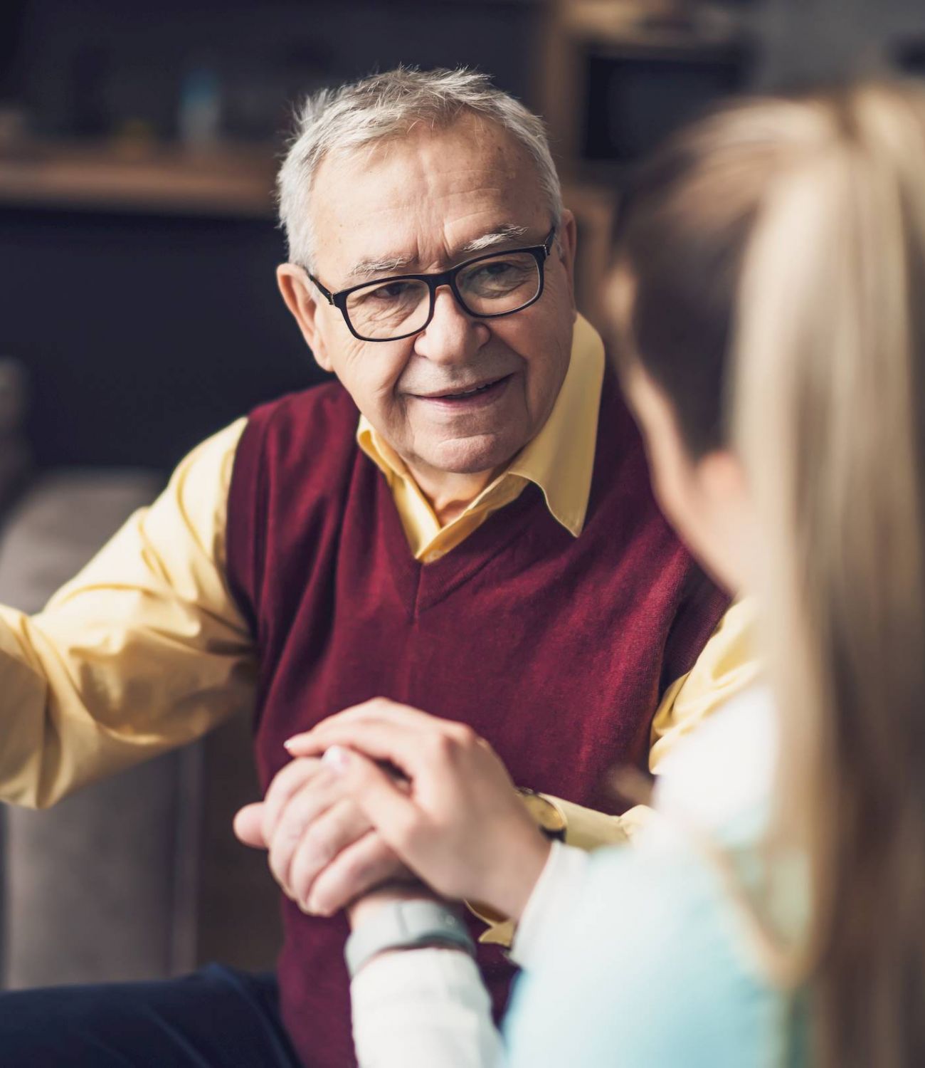 An elderly man with glasses smiles while holding hands with a younger woman in the cozy living room at Barton House.