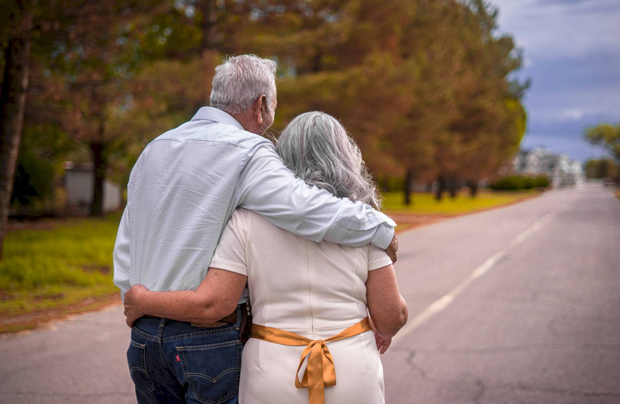 An elderly couple walks arm in arm down a tree-lined road, Honoring Every Story on a cloudy day.