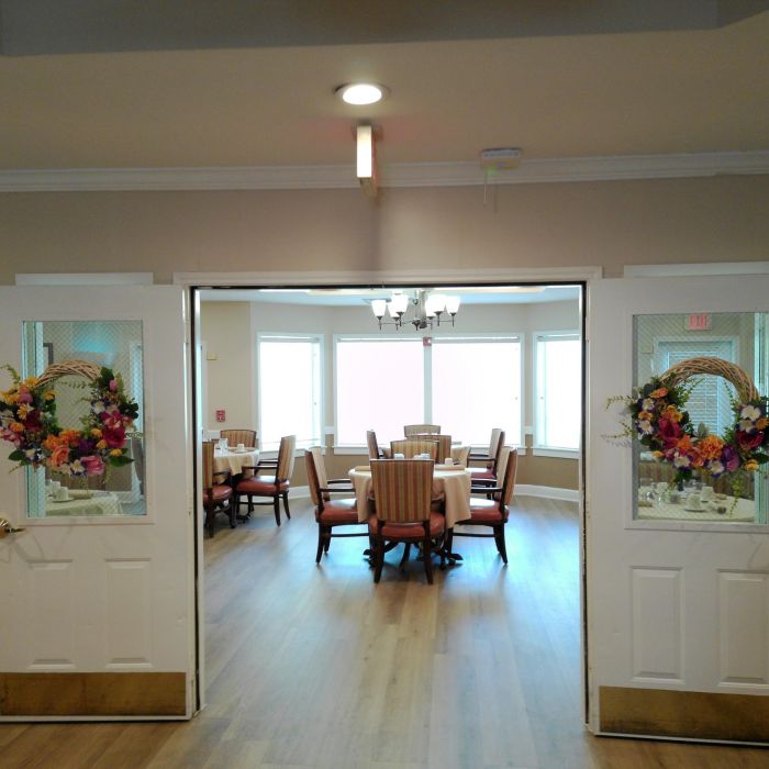 Double doors with floral wreaths open to a bright dining room at Tarina of Kingwood, featuring tables, chairs, and large windows.