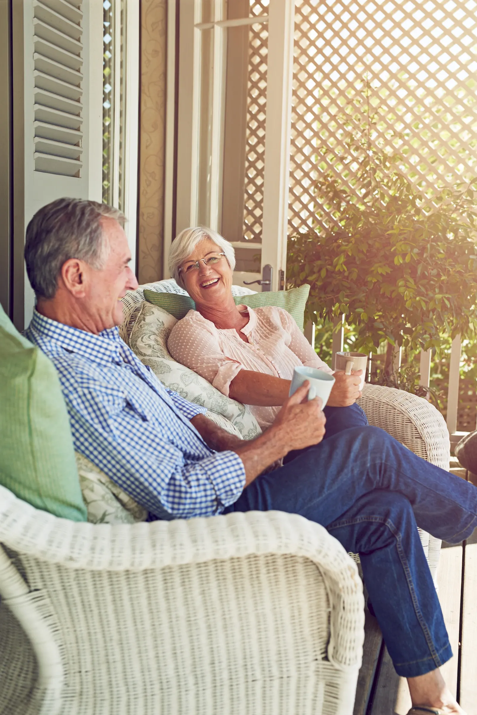 Smiling older couple sitting on a porch with coffee mugs, enjoying a relaxed conversation together.