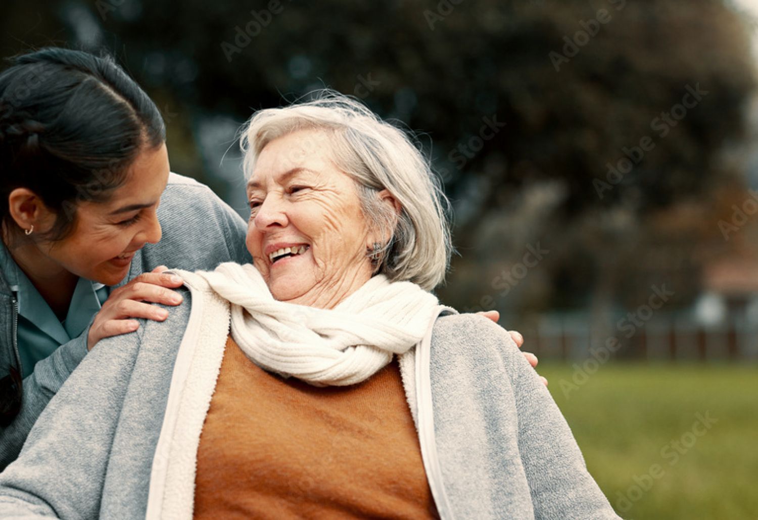 A young woman embraces and smiles at an elderly woman in a scarf, both enjoying time together outdoors.