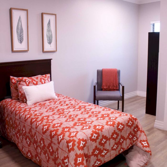 A neatly made bed with a red patterned comforter, a chair with a red throw, and two leaf prints on the wall.