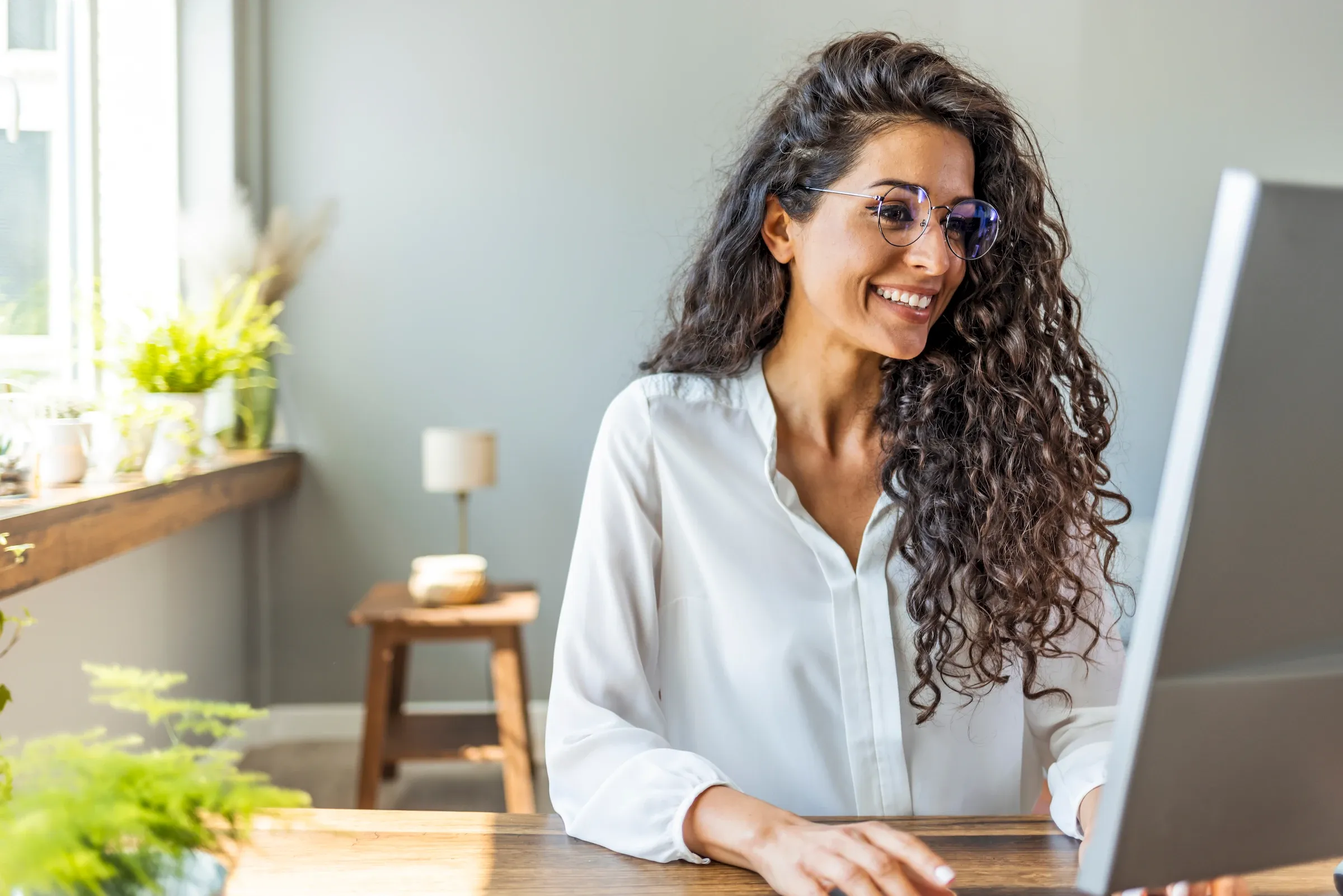 Smiling woman with curly hair and glasses working at a computer in a bright, modern home office.