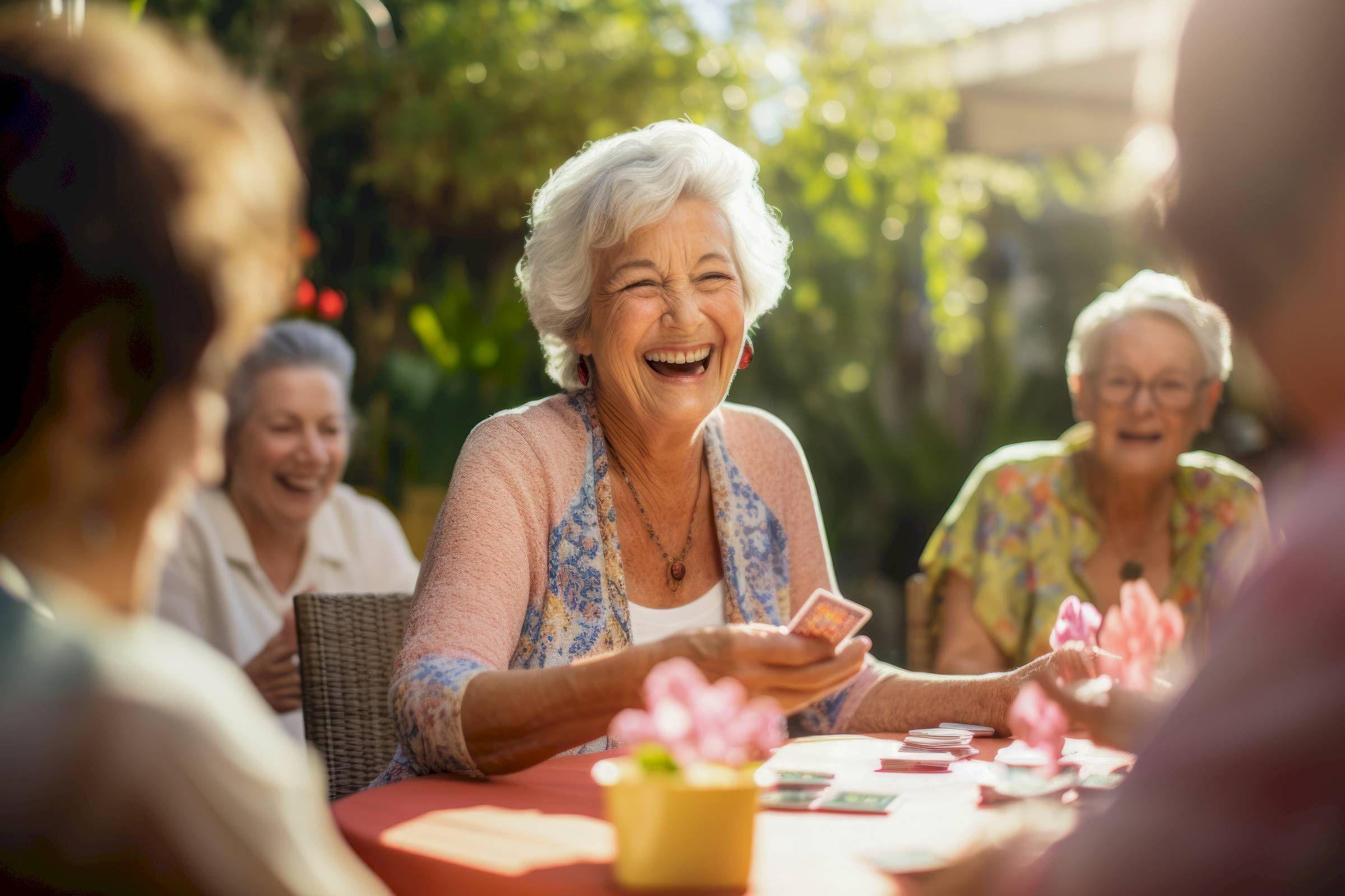 Four elderly women, including Tarina of Stockton, smile and play cards at a sunlit outdoor table, surrounded by plants and flowers.