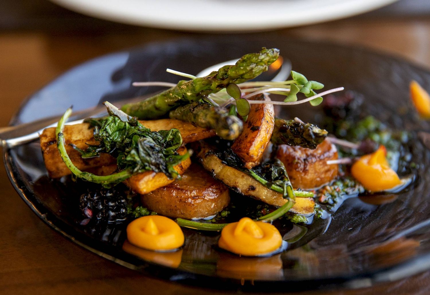 Gourmet roasted vegetables, asparagus, and greens on a black plate with orange sauce dollops.