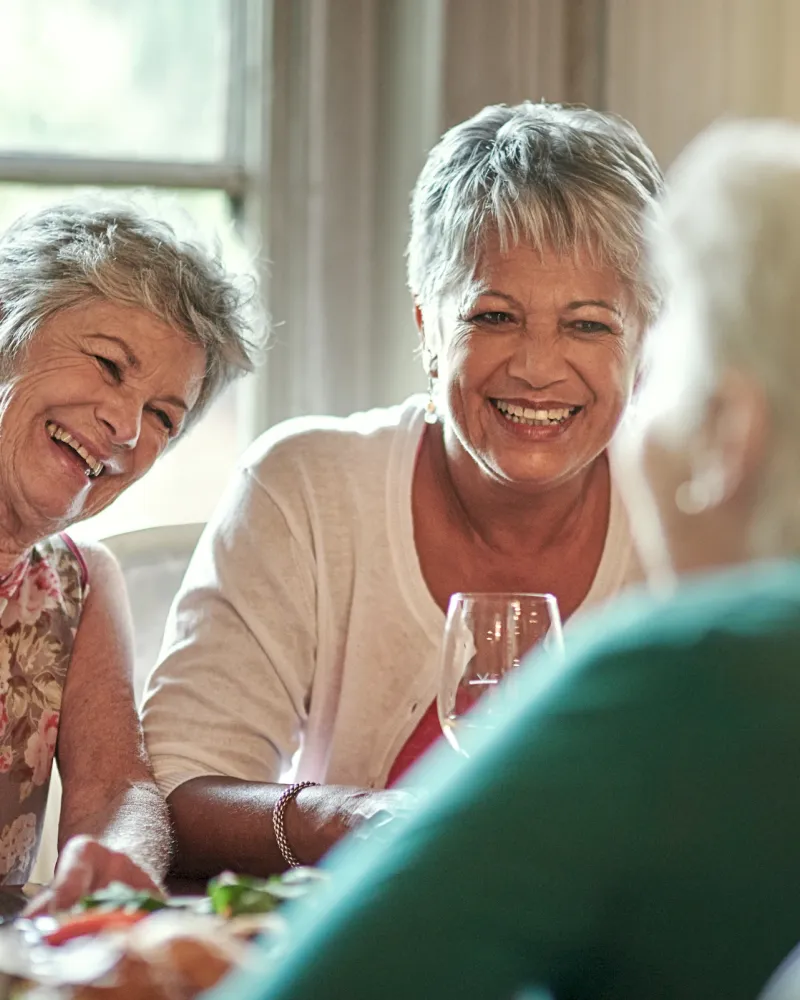 Three older women laughing and enjoying wine together at a dining table in a sunlit room.