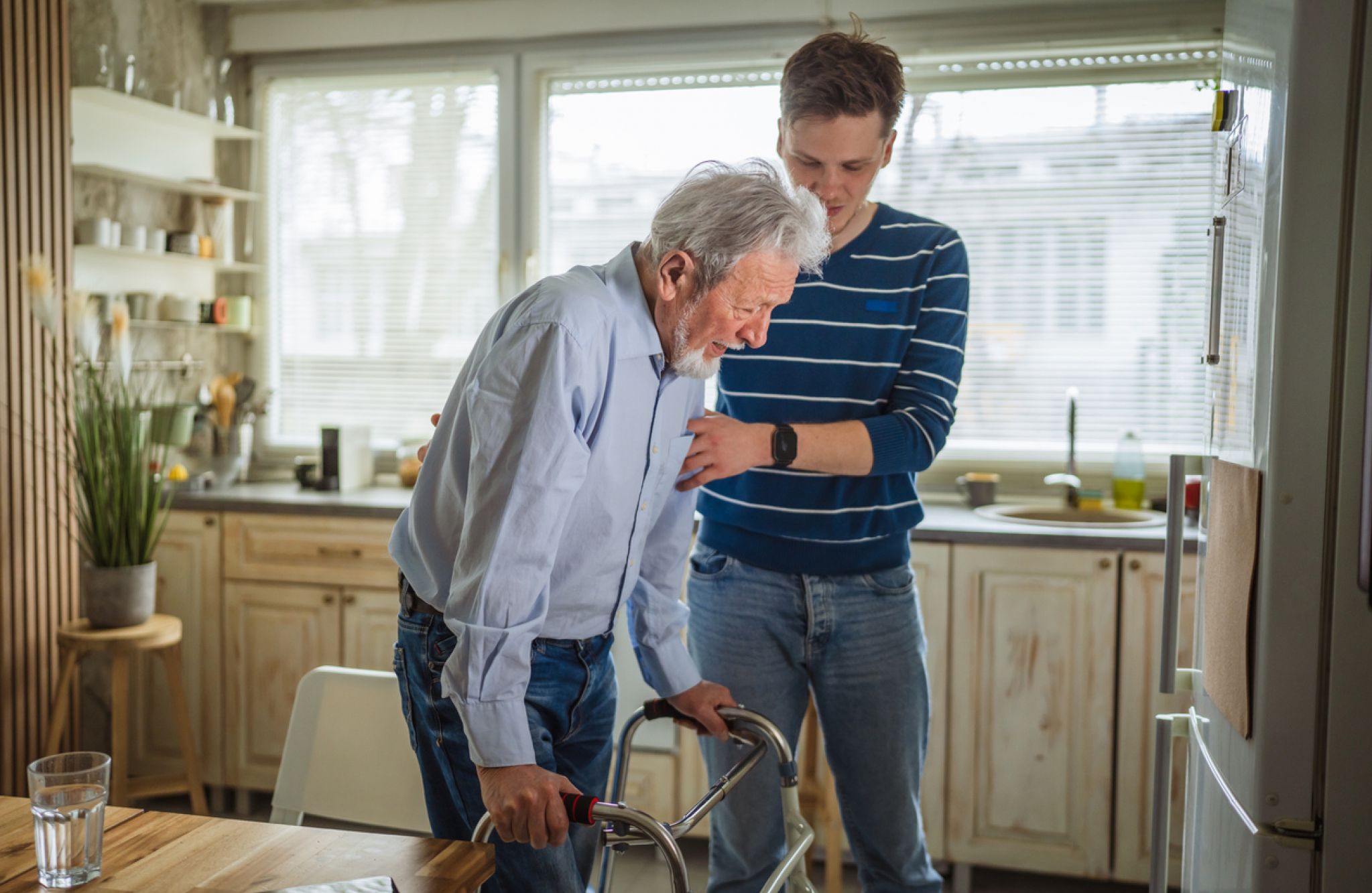 Young man helps elderly man with a walker in a bright kitchen, discussing signs of assisted living and future care options.