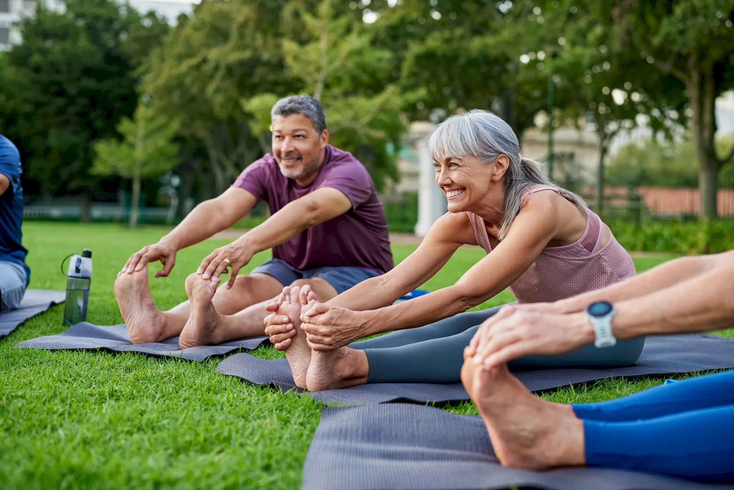 Older adults stretch on yoga mats outdoors, smiling and reaching for their feet in a group exercise session.