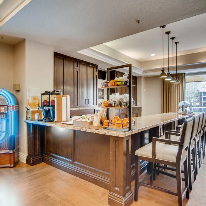 The Terraces at Kingwood offers a modern hotel breakfast bar with a jukebox, drinks, fruit, and high chairs by large windows for senior living.