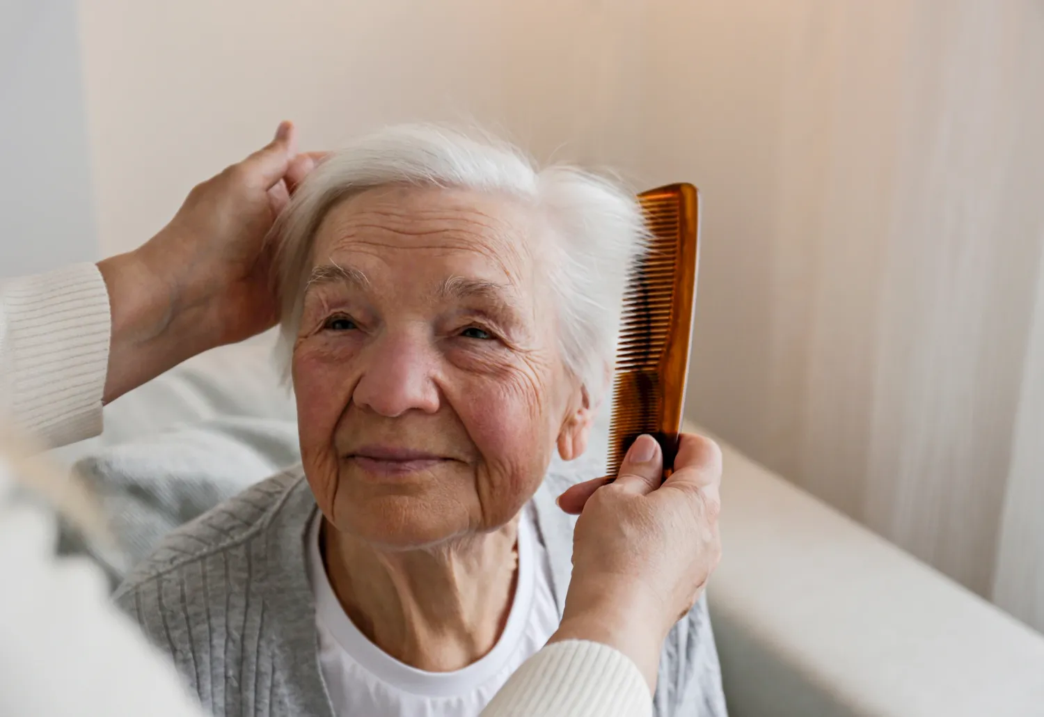 An elderly woman smiles while someone gently combs her white hair with an amber-colored comb.