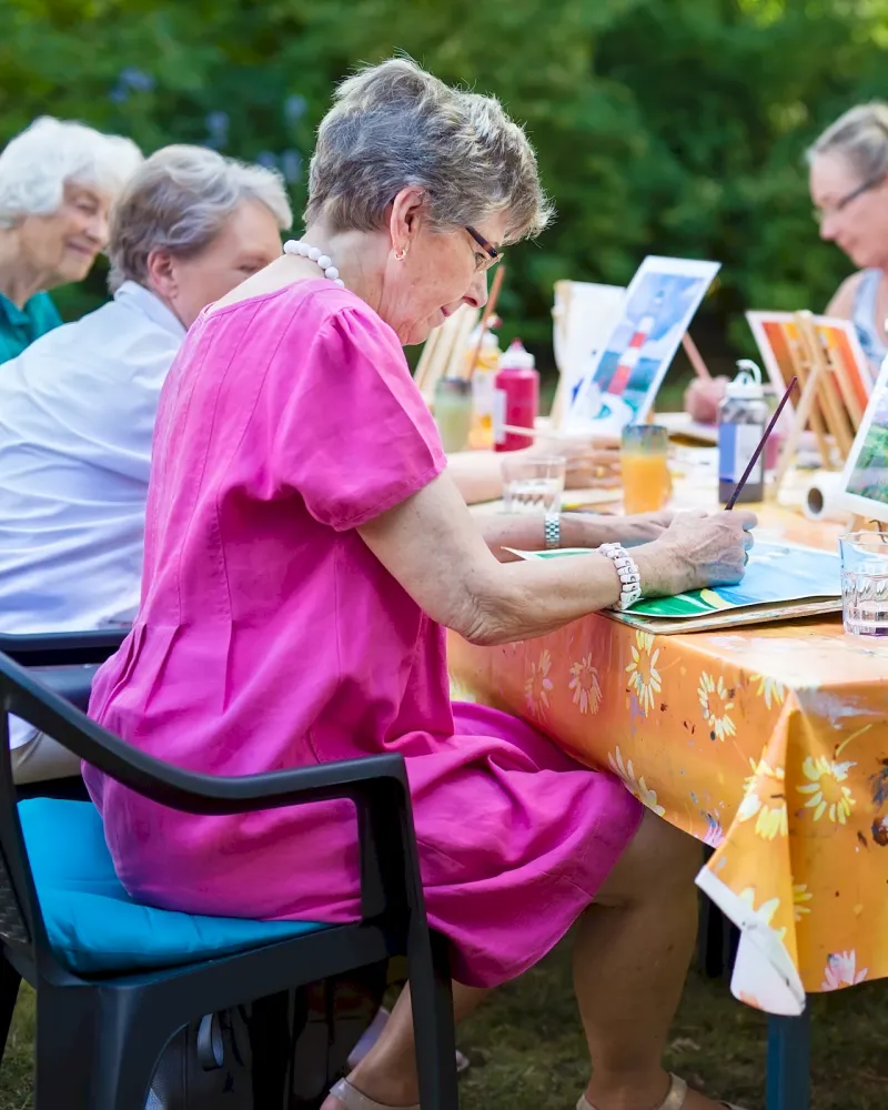 Six older women painting together outdoors at a table covered with a floral tablecloth on a sunny day.