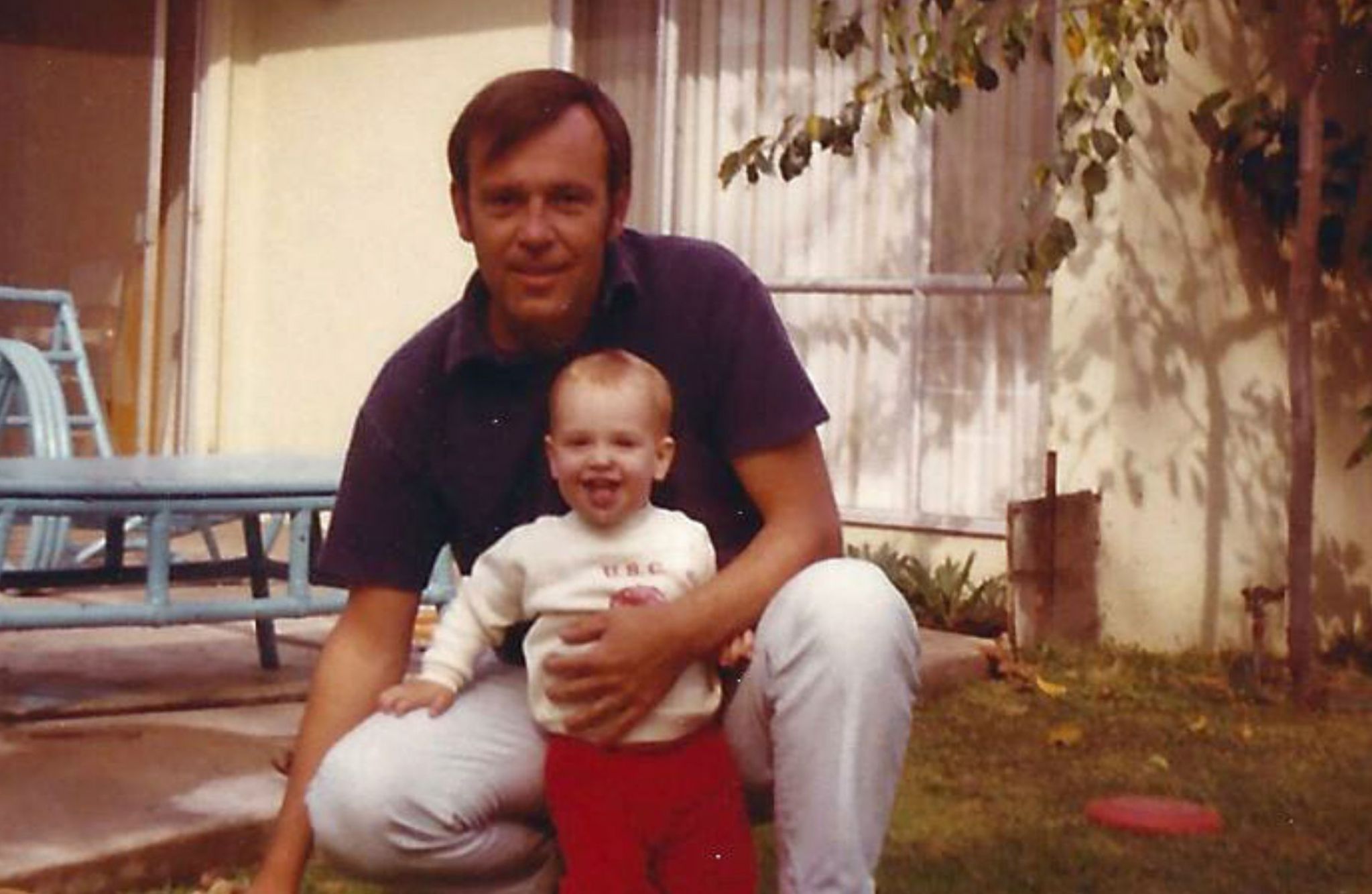 An adult crouches, hugging a smiling baby in a yard—Honoring Every Story amid patio furniture and the home's warm background.