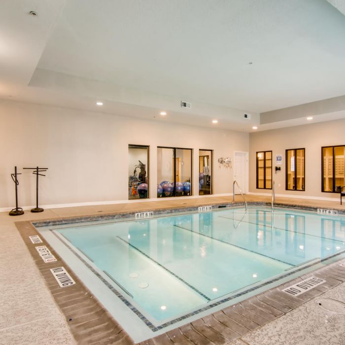 Indoor swimming pool with clear water at The Terraces at Kingwood, featuring chairs and gym equipment behind glass doors.