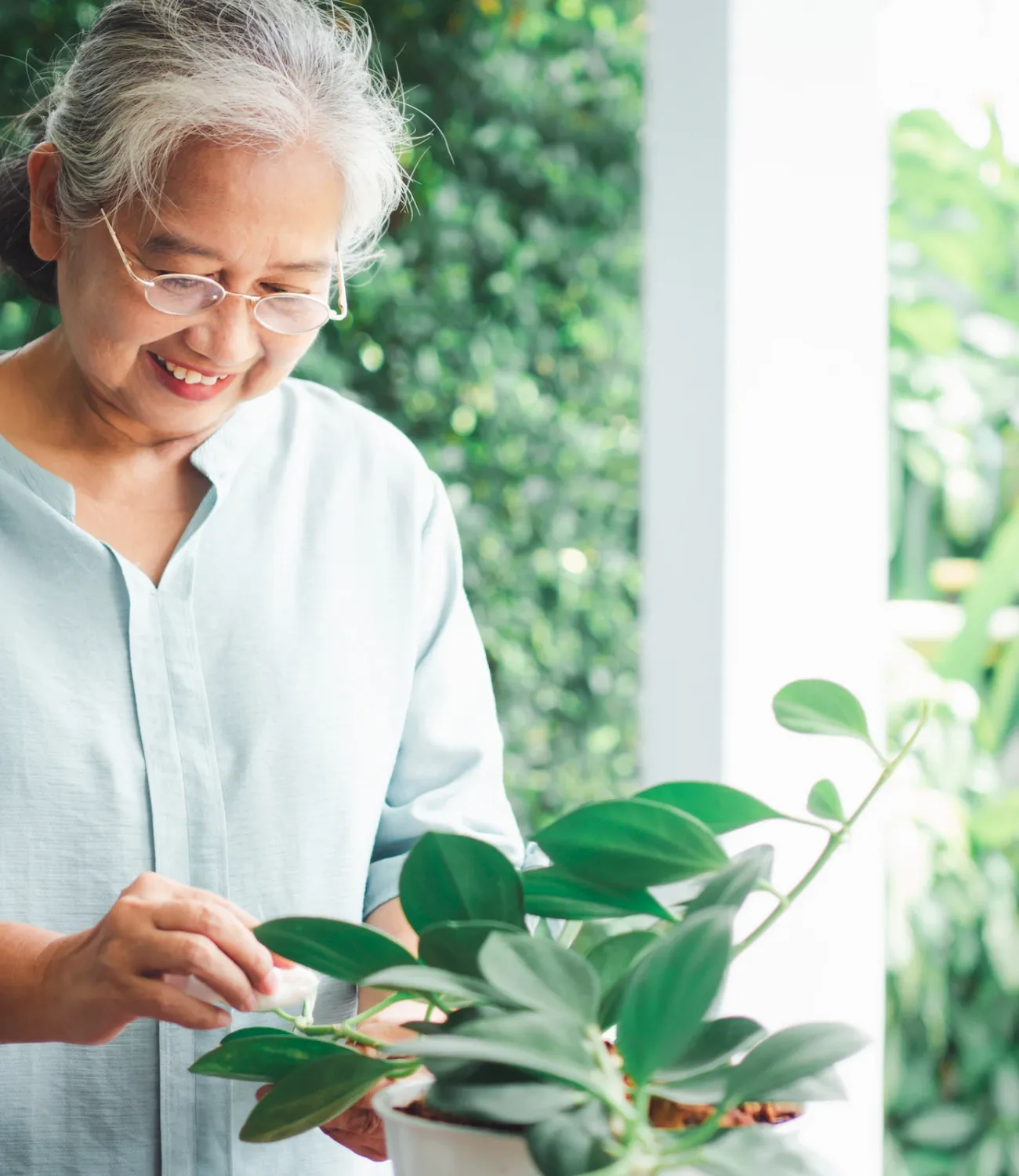 Smiling older woman wearing glasses tending to a potted plant outdoors surrounded by greenery.