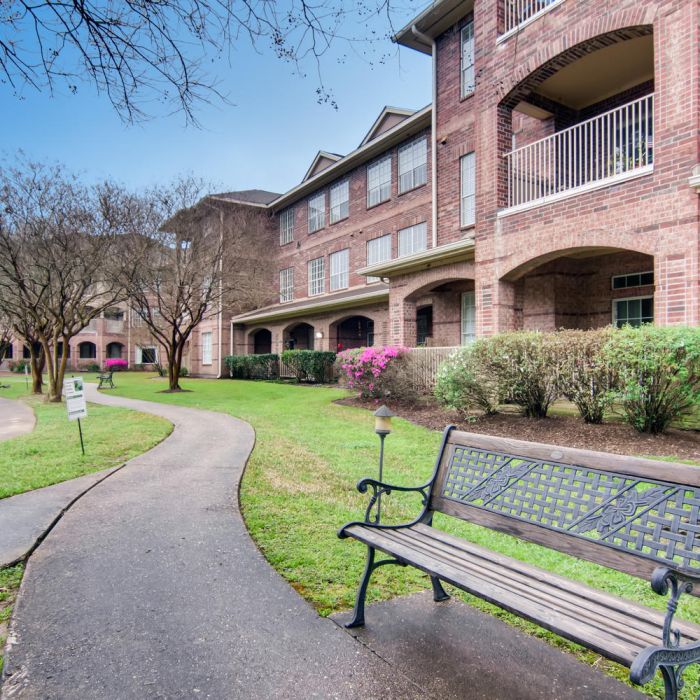 A curved sidewalk, bench, and pond next to Terraces at Kingwood senior living apartments with balconies in Kingwood TX.